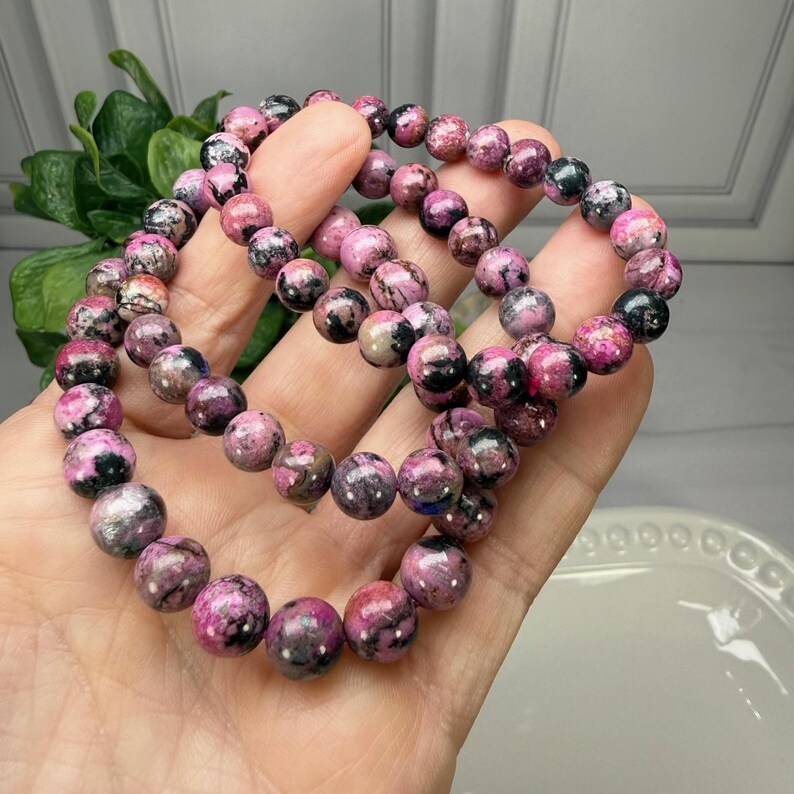 May include: Two beaded bracelets with pink and black stones. The bracelets are made of round, polished beads in varying shades of pink and black. The bracelets are held in a hand, showcasing the color and texture of the stones.