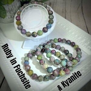 May include: Two bracelets made of colorful gemstone beads. The bracelets are layered on a white surface. The text "Ruby in Fuchsite & Kyanite" is written on the surface.