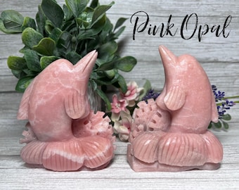 May include: Two pink opal dolphin sculptures with a floral design base. The sculptures are carved from pink opal and have a smooth, polished finish. The text "Pink Opal" is written in black cursive lettering at the top of the image.