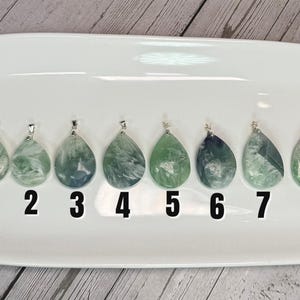May include: Eight teardrop-shaped pendants with silver bails, each showing unique patterns of green, blue, and white within the stone. The pendants are numbered 1 to 8 and are displayed on a white, oval-shaped tray.