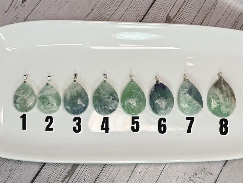 May include: Eight teardrop-shaped pendants with silver bails, each showing unique patterns of green, blue, and white within the stone. The pendants are numbered 1 to 8 and are displayed on a white, oval-shaped tray.