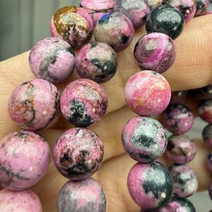 May include: Close-up of a bracelet featuring round, polished beads. The beads are primarily pink with dark gray and black marbling. The bracelet is strung together, showcasing the natural variations in color and pattern of the gemstones.
