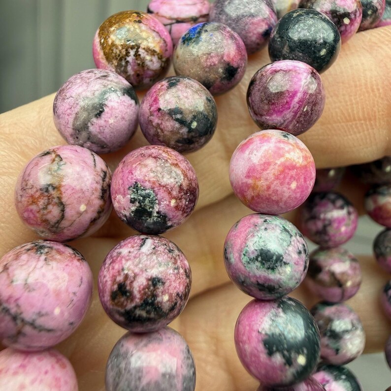 May include: Close-up of a bracelet featuring round, polished beads. The beads are primarily pink with dark gray and black marbling. The bracelet is strung together, showcasing the natural variations in color and pattern of the gemstones.