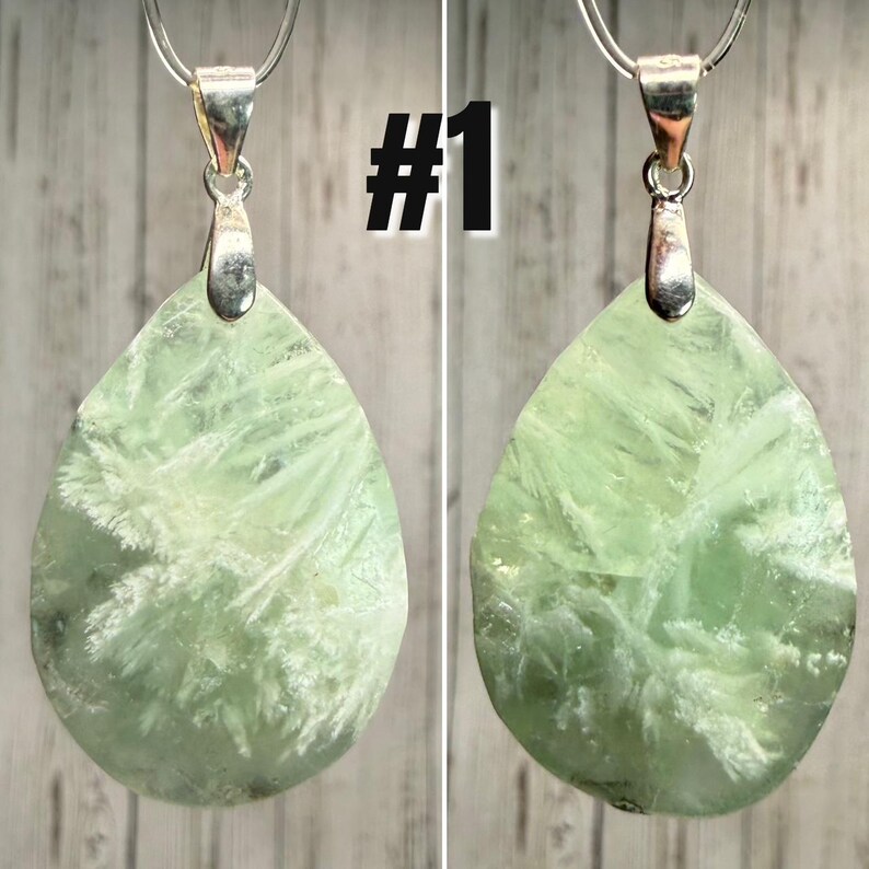May include: Two pendants featuring a light green, teardrop-shaped stone with white, feathery inclusions. Each stone has a silver-coloured bail. The pendants are displayed side-by-side. The text "#1" is in the upper right corner.