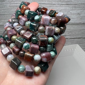 May include: A stack of bracelets made with green, brown, and purple gemstone beads. The beads are square and round in shape.