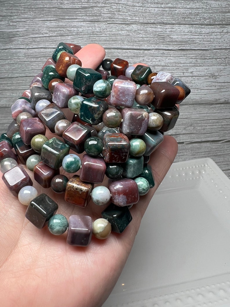 May include: A stack of bracelets made with green, brown, and purple gemstone beads. The beads are square and round in shape.
