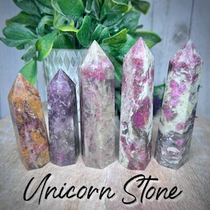 May include: Five polished unicorn stone points in various shades of pink, purple, and white. The points are arranged in a row on a wooden surface.  Unicorn Stone.