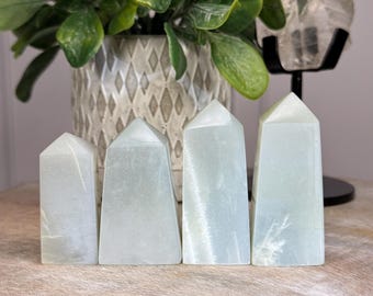 May include: Four pale green crystal obelisks of varying heights. Each obelisk has a rectangular base and a pointed top. The crystals have a translucent quality, with subtle internal patterns. The obelisks are arranged in a row, suggesting a collection or set.