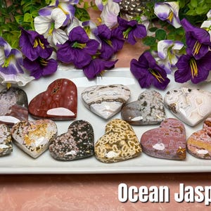 May include: A collection of heart-shaped Ocean Jasper stones in various colors, including red, white, brown, and pink, arranged on a white rectangular plate. The text "Ocean Jasper" is at the bottom. Purple and white flowers are in the background.