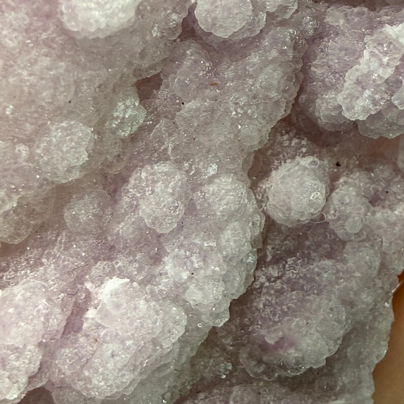 May include: Close-up of a crystalline mineral formation. The image showcases a light purple and white textured surface with rounded, clustered formations. The overall appearance suggests a natural, geological specimen, possibly a geode or crystal cluster.