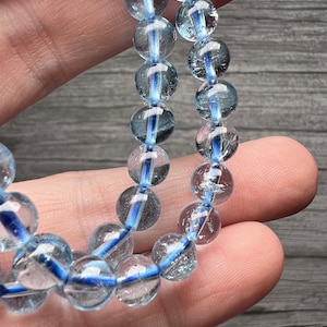 May include: A close-up of a bracelet made of clear, round beads with a blue stripe running through each bead. The bracelet is strung on a blue elastic cord. The beads are held in a hand.
