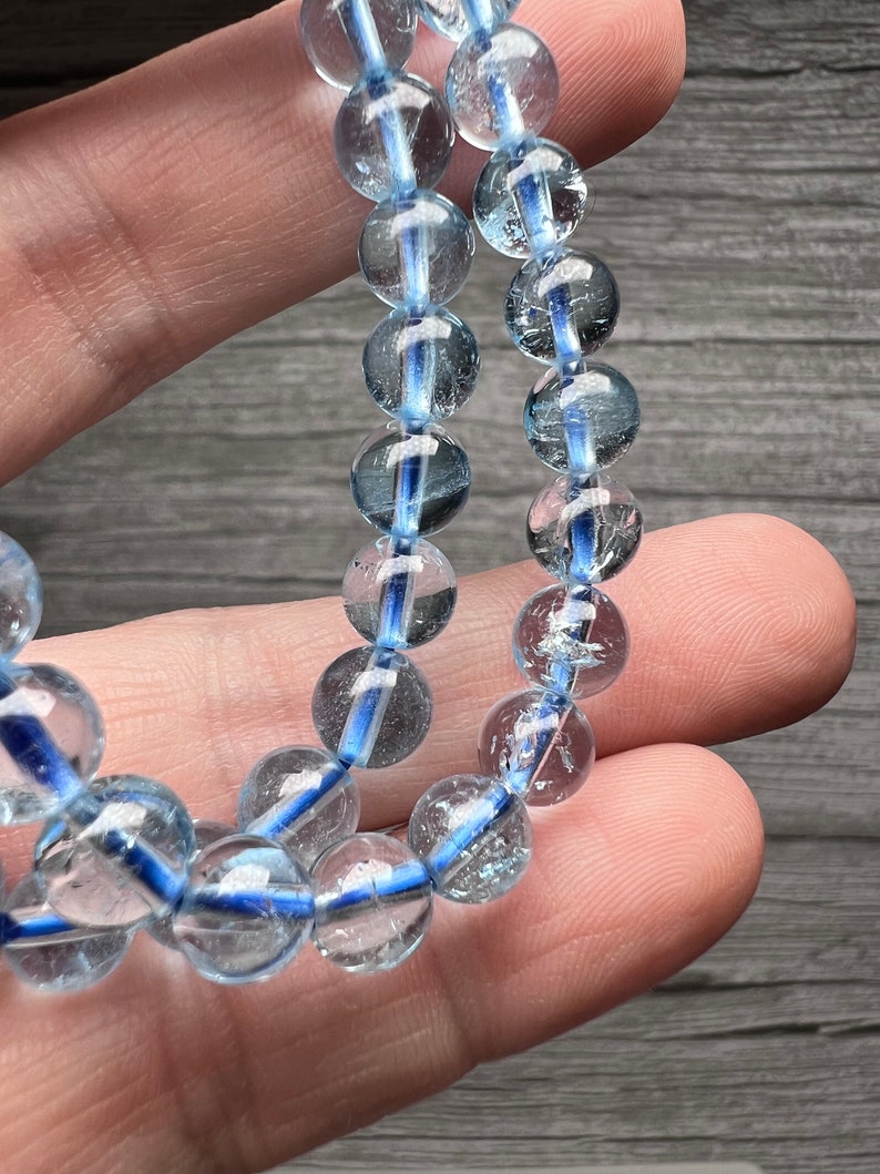 May include: A close-up of a bracelet made of clear, round beads with a blue stripe running through each bead. The bracelet is strung on a blue elastic cord. The beads are held in a hand.