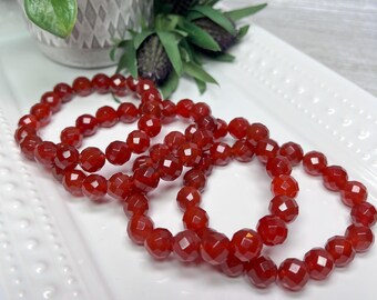 May include: Three red carnelian gemstone bracelets with faceted beads. The bracelets are stacked on a white surface.