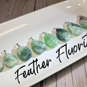 May include: A selection of teardrop-shaped fluorite pendants in shades of green and white, each with a silver-coloured bail. The pendants are displayed on a white rectangular tray. The words "Feather Fluorite" are written in black script.