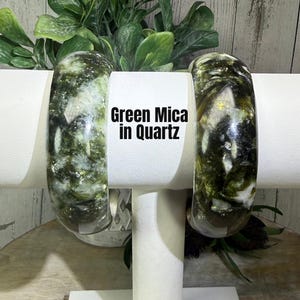 May include: Two Green Mica in Quartz bangles are displayed on a white stand. The bangles have a marbled appearance with green and white swirls. The text "Green Mica in Quartz" is visible on the stand. The background includes green foliage.