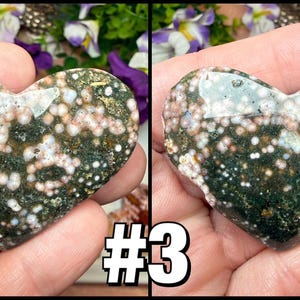 May include: A heart-shaped stone with a speckled pattern of white, pink, and green. The polished surface of the stone reflects light, and the shape is held in a hand. The number "3" is visible in the lower right corner.