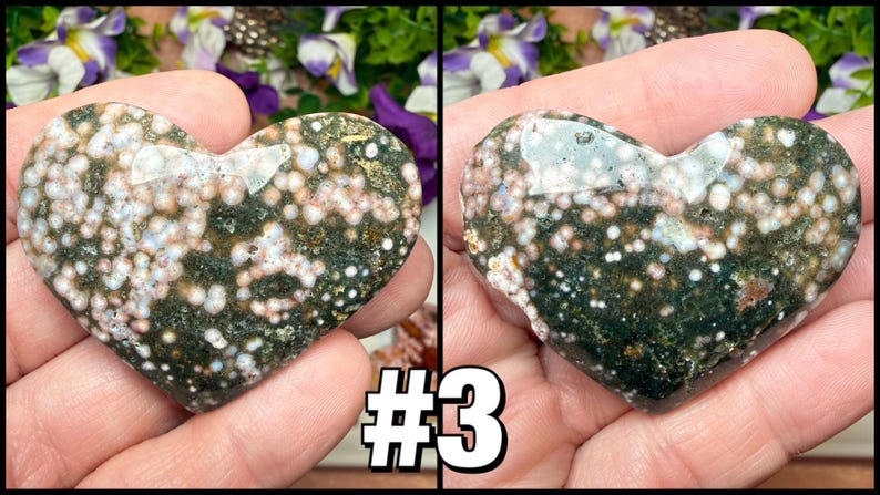 May include: A heart-shaped stone with a speckled pattern of white, pink, and green. The polished surface of the stone reflects light, and the shape is held in a hand. The number "3" is visible in the lower right corner.