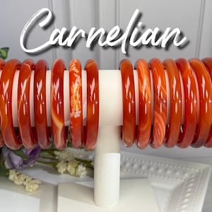 May include: A collection of smooth, polished carnelian bangles displayed on a white T-bar stand. The bracelets feature varying shades of translucent orange and deep red, with some showing natural white banding and swirling patterns throughout.
