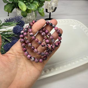 May include: Three beaded bracelets with pink and black stones. The bracelets are held in a hand, and are resting on a white, oval-shaped tray. The beads are round and appear to be approximately 0.3 inches in diameter. The background includes greenery and a crystal.