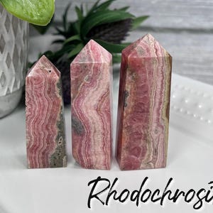 May include: Three pink and white rhodochrosite crystal points, also known as "the stone of compassion", are arranged in a row on a white surface. The word "Rhodochrosite" is written in black below the crystals.