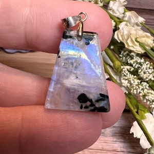 May include: A silver pendant with a triangular iridescent moonstone with black inclusions. The pendant is held in a hand.