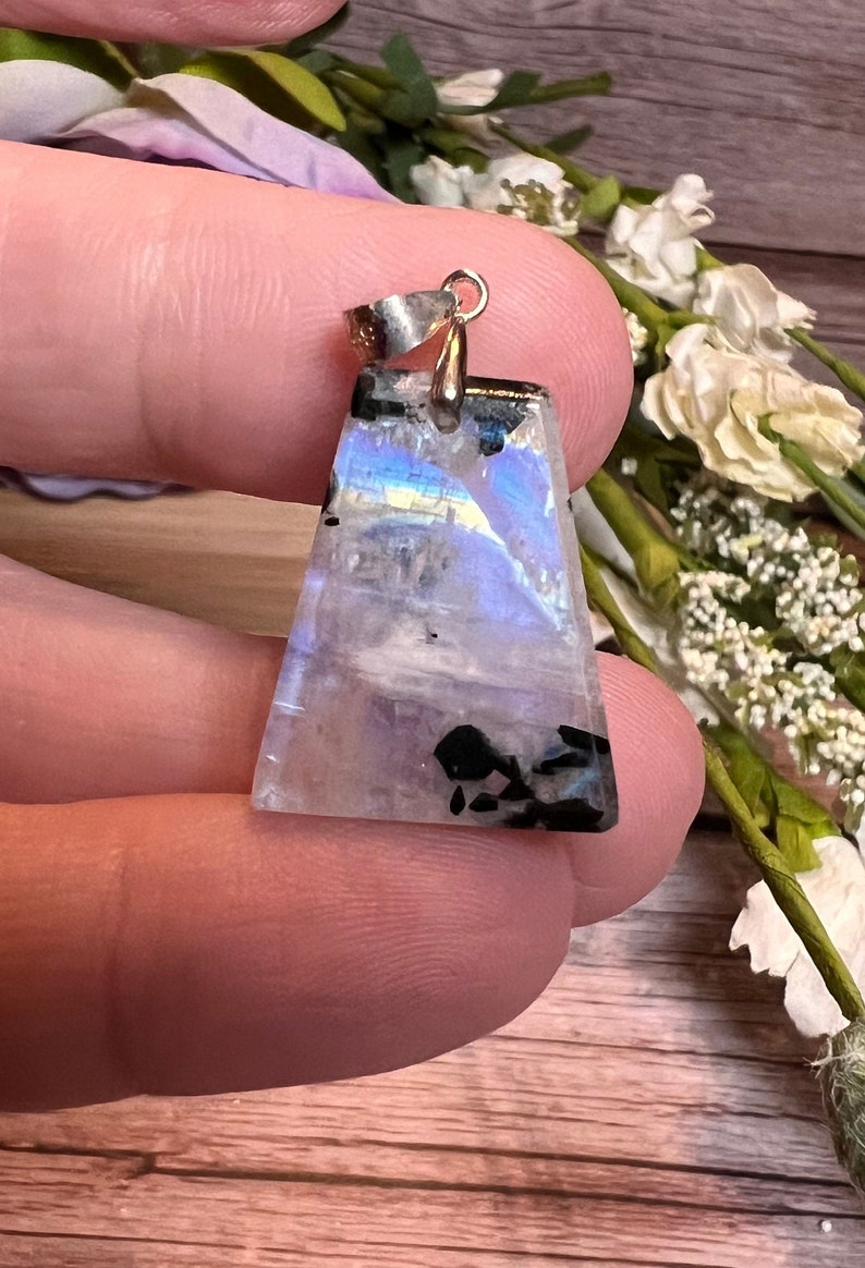 May include: A silver pendant with a triangular iridescent moonstone with black inclusions. The pendant is held in a hand.