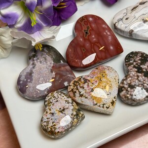 May include: A collection of heart-shaped stones in various colors and patterns, including red, purple, and speckled designs. The stones are arranged on a white surface, with purple flowers in the background. These decorative stones are ideal for home decor or as a unique gift.