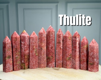 May include: A collection of eleven polished Thulite crystal towers arranged in a row. Each stone features a mottled texture with varying shades of pink, red, and cream-colored inclusions. The word Thulite is printed in white text in the upper right.