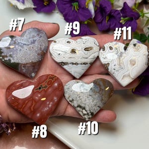 May include: Five heart-shaped polished stones in various colors and patterns. The stones are numbered #7, #8, #9, #10, and #11. The stones are held in a hand, with purple flowers in the background. The stones are made of different types of minerals.