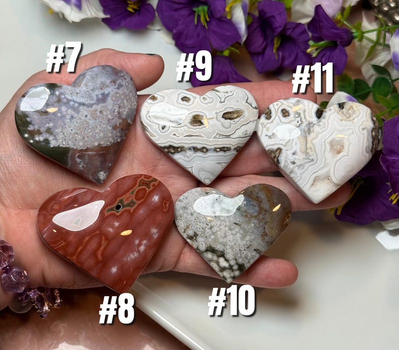 May include: Five heart-shaped polished stones in various colors and patterns. The stones are numbered #7, #8, #9, #10, and #11. The stones are held in a hand, with purple flowers in the background. The stones are made of different types of minerals.