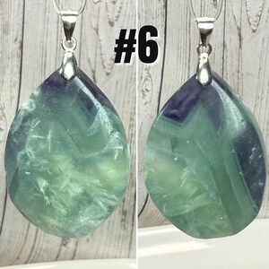 May include: A pendant with a silver bail, showcasing a teardrop-shaped, translucent stone in green and purple hues. The stone has a crystalline texture. The image includes the number "#6" in black on the top right.