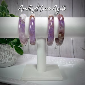 May include: Four amethyst lace agate bangles in shades of purple and white on a white display stand. The bangles are stacked on top of each other. Amethyst Lace Agate is written in black text above the bangles.