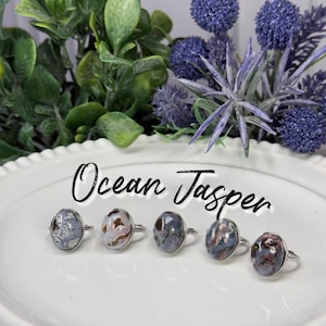 May include: Five silver-toned rings featuring oval-shaped Ocean Jasper gemstones with varied patterns of gray, blue, and brown. The text Ocean Jasper is written in black script above the rings, which are arranged on a white scalloped plate.