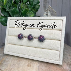 May include: Three silver rings with ruby in kyanite gemstones. The rings are displayed on a white fabric ring display with the text "Ruby in Kyanite" written on the top.