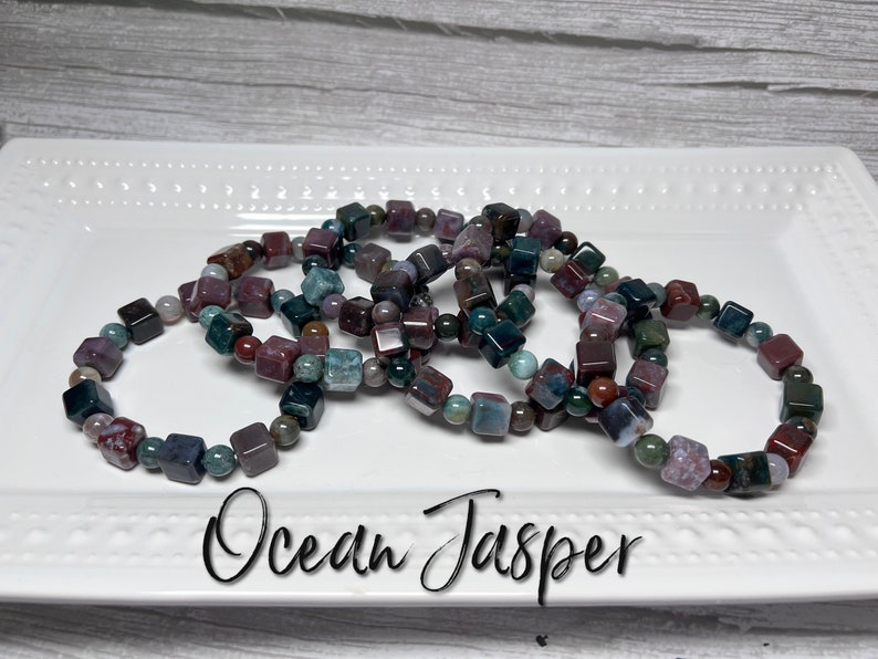 May include: A set of five bracelets made with ocean jasper beads. The beads are a mix of green, brown, and red colors. The bracelets are stacked on a white plate with the words "Ocean Jasper" written below them.