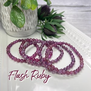 May include: A set of five pink and purple gemstone bracelets, stacked on top of each other. The bracelets are made of small, round beads. The text "Flash Ruby" is written in pink and purple letters below the bracelets.