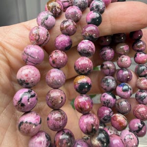 May include: Close-up of a strand of round, polished beads. The beads are primarily a vibrant pink color, with darker pink and black marbling throughout. The beads are approximately 1 cm in diameter, strung together to form a necklace or bracelet.