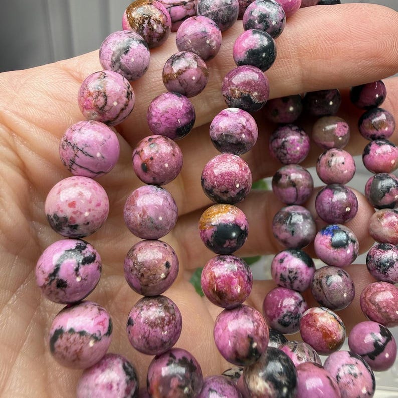 May include: Close-up of a strand of round, polished beads. The beads are primarily a vibrant pink color, with darker pink and black marbling throughout. The beads are approximately 1 cm in diameter, strung together to form a necklace or bracelet.