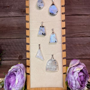 May include: Six iridescent moonstone pendants on display on a wooden stand. The pendants are various shapes and sizes, including teardrop, triangle, and oval. The moonstone is a milky white color with flashes of blue and grey.