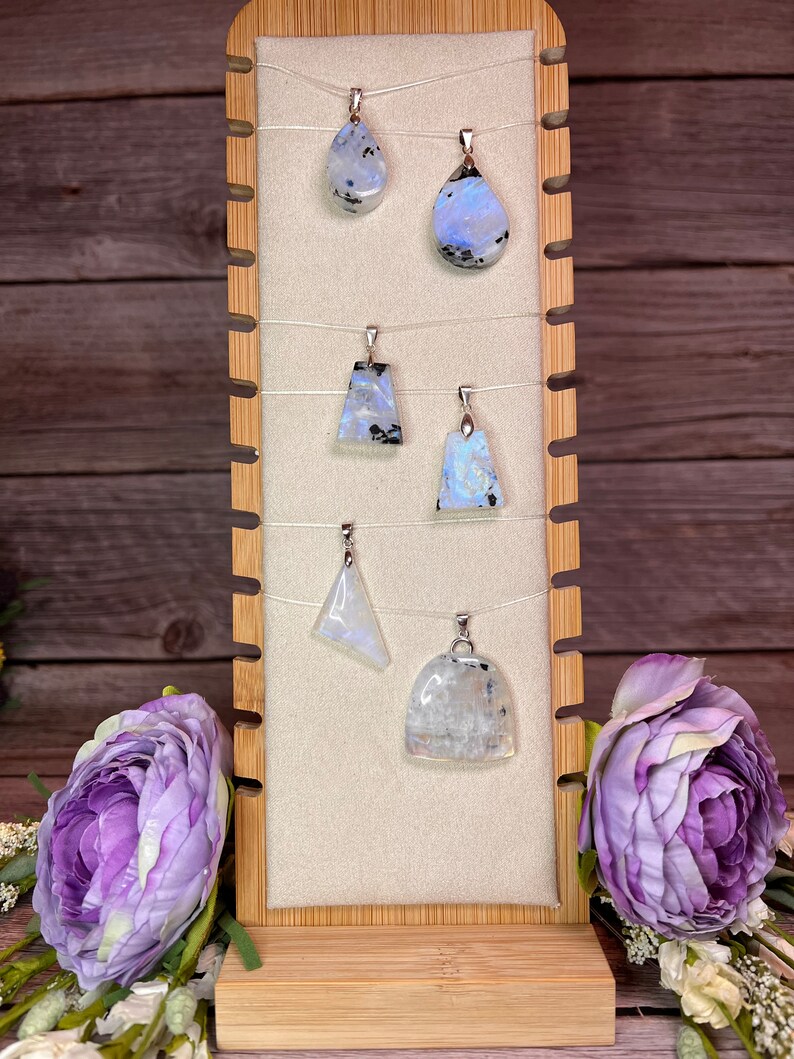 May include: Six iridescent moonstone pendants on display on a wooden stand. The pendants are various shapes and sizes, including teardrop, triangle, and oval. The moonstone is a milky white color with flashes of blue and grey.