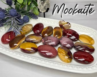 May include: A collection of polished Mookaite stones in shades of red, yellow, and brown, arranged on a white, oval-shaped platter. The stones vary in size and shape, with a heart-shaped stone included. The word "Mookaite" is written in cursive.