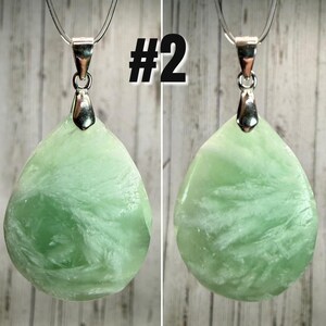 May include: Two pendants featuring a light green, teardrop-shaped stone with white veining. Each stone has a silver-coloured bail. The pendants are displayed against a light wood-grain background. The number "#2" is printed in black.