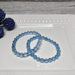 May include: Two light blue beaded bracelets are displayed on a white rectangular plate. The bracelets are made of small, round, translucent beads strung on a blue elastic cord. Decorative blue floral accents are in the background.