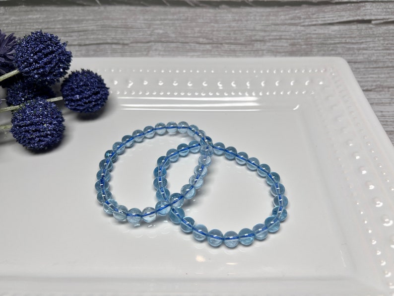 May include: Two light blue beaded bracelets are displayed on a white rectangular plate. The bracelets are made of small, round, translucent beads strung on a blue elastic cord. Decorative blue floral accents are in the background.