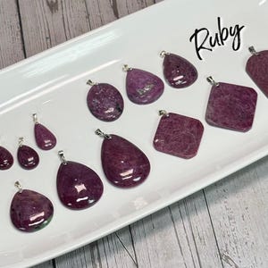 May include: A collection of ruby pendants in various shapes, including teardrop, oval, and square, displayed on a white rectangular tray. Each pendant has a silver bail. The word "Ruby" is written in cursive in the upper right corner.