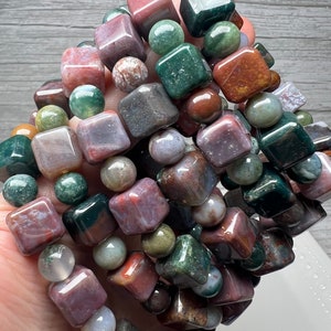 May include: A close-up of a stack of bracelets made with green, brown, and pink agate gemstone beads. The beads are square and round in shape.