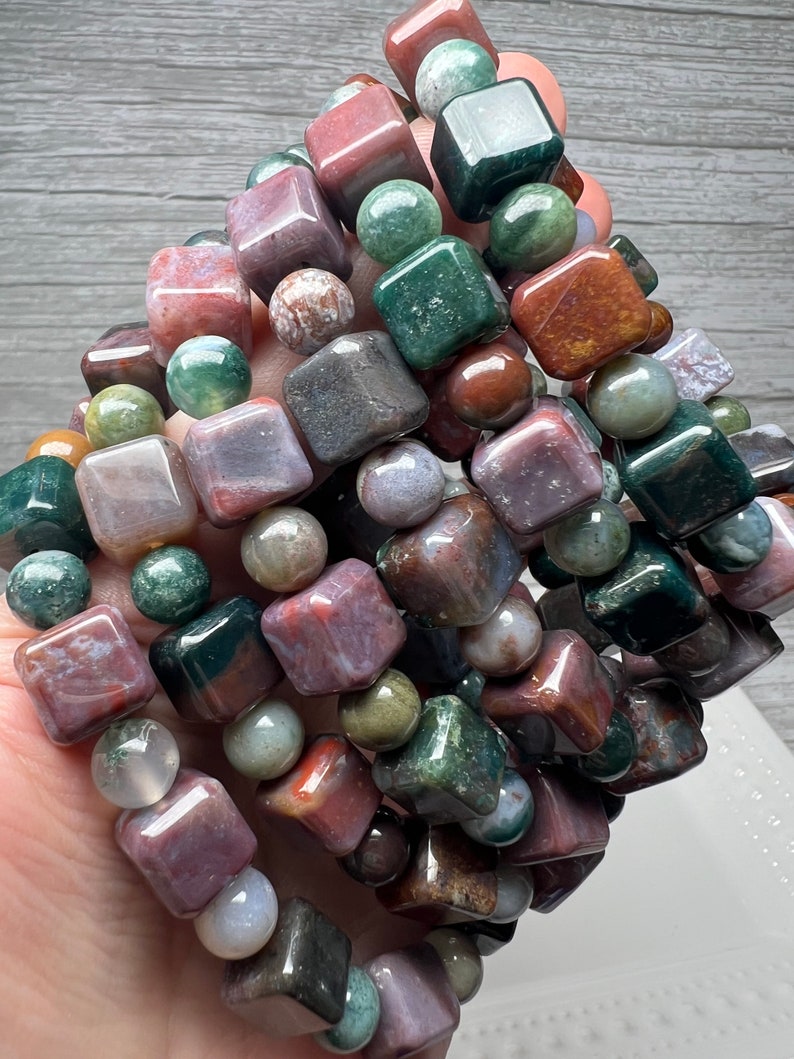 May include: A close-up of a stack of bracelets made with green, brown, and pink agate gemstone beads. The beads are square and round in shape.