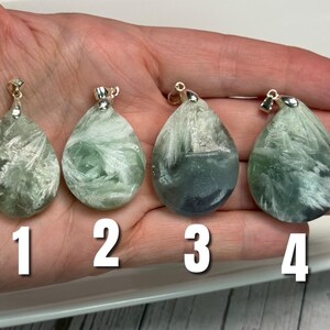 May include: Four teardrop-shaped pendants with silver-coloured bails. The pendants are a mix of green and white, with a feathery, textured appearance. Each pendant is numbered 1 through 4 in white. The pendants are displayed on a hand.