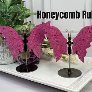 May include: Two butterfly-shaped ornaments with textured, deep pink wings and black bodies, each mounted on a thin metal rod with a circular base. The text "Honeycomb Ruby" is displayed in black letters above the butterfly figures.