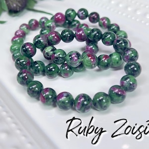 May include: A bracelet made of green and pink ruby zoisite beads. The bracelet is made of multiple strands of beads and is stacked on a white surface. The text "Ruby Zoisite" is written in black below the bracelet.